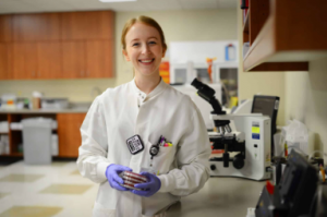 A young woman in a laboratory with red hair pulled back, wearing a white lab coat with purple gloves.  She is smiling. There is a microscope behind her and the background is slightly blurry.