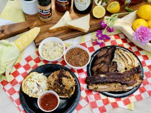 An Aerial Shot of a Hot Spot Picnic Spread featuring brisket, ribs, and sliced smoked chicken breast on one plate. Another plate shows a pulled pork sandwich with coleslaw on it and Hot Spot's signature sauce. Two of their side dishes are posed to the sid