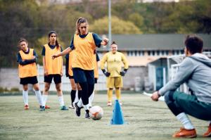A female soccer player demonstrates ball control skills amidst a line of cones during rigorous training. While teammates await their turns, the coach offers guidance from the sidelines.