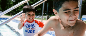 Two boys are playing and splashing in a swimming pool, with one wearing a "Cool Dude" shirt and the other squinting as water splashes his face.