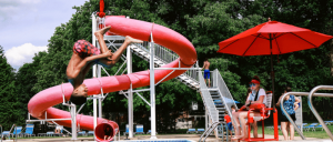 A boy is flipping into a swimming pool while a lifeguard watches nearby, with a red water slide in the background.