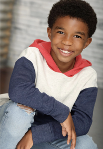 Portrait of a young African American boy wearing a casual multicolored hoodie and jeans, smiling warmly against a gray textured background. Showcasing the vibrant youth talent at The Industry Network.