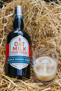 Photo of a spirits bottle, nestled in a bed of straw, with a rocks glass with ice next to it holding a light-brown cream liqueur, Hansen's Oat Milk Cream Liqueur Spirit.