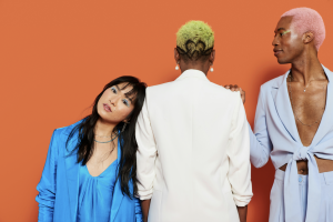 Three people posing in front of orange backdrop: Person with long wavy dark hair and fringe facing the camera, next person with short green-dyed hair facing the back, then person with short pink-dyed hair facing the person in the middle