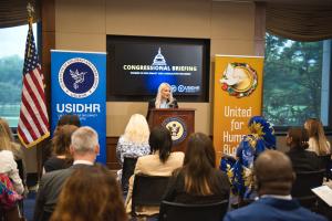 A blonde woman, Dr. Isabelle Vladoiu, speaks at a podium during a congressional briefing on women in diplomacy, with attendees and banners in the background.