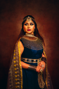 An image of a woman dressed as Cleopatra in elaborate blue and gold Egyptian attire. She is adorned jewelry and a headdress. She looks regal and authoritative and evokes the grandeur and mystique of ancient Egypt.