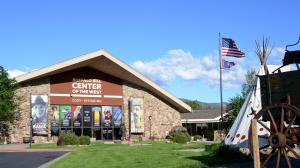 The facade of the Buffalo Bill Center of the West on the left with an American flag, and partial view of a white teepee and chuckwagon on the right