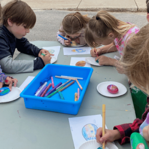 Children drawing on paper at outdoor table.