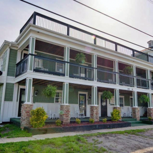 Photo of the green and white two story Carolina Beach Inn. Rocking chairs sit on the front and there is a second floor covered veranda.