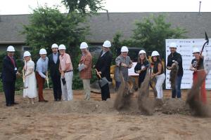 A groundbreaking ceremony with a group of people standing behind a dirt pile holding up shovels of dirt and smiling.