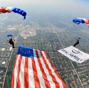 Two parachuters in the air with red white and blue parachutes holding an american flag and SEAL Family banner.