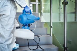 Close-up of a disinfection technician in full PPE holding a blue and gray disinfectant sprayer, actively disinfecting a staircase with glass balustrades.