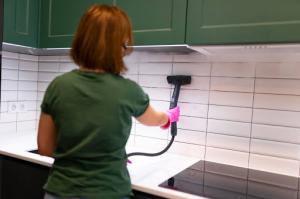 A woman in a green shirt and pink latex gloves uses a black steam cleaner to clean kitchen tiles with green cupboards in the background.