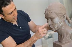 Dr. Simon Ourian, wearing a dark t-shirt, is seen sculpting a bust. He is carefully working on the facial features of the sculpture, highlighting his artistic talent and meticulous approach.