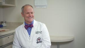 photo of Jerry Grimes, M.D., smiling as he sits in a medical exam room. He is wearing a blue button down shirt and purple bow tie with small white polka dots under his medical white coat.
