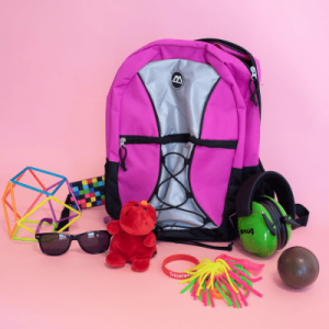 A pink and grey backpack is surrounded by various items, including a colorful geometric toy, a multicolored cube, black sunglasses, a small red plush toy, green noise-canceling headphones, a Triceratops bracelet, a neon string toy, and a small ball.