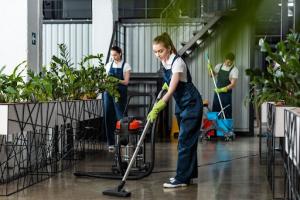 Three commercial cleaners in white shirts, navy blue overalls, and black sneakers are actively dusting, vacuuming, and mopping the floors of an office. The office space is adorned with plants in plant boxes on black steel stands. In the background, a blac
