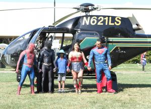 Spiderman, Batman, a child, Wonder Woman, and Superman hold hands in front of the black and green helicopter.
