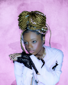 This image features Seedel, a poet & mental health advocate, posing against a pink background. With intricate braids & a pink shirt paired with black lace gloves, Seedel radiates style and confidence.her contributions to literature & social causeles