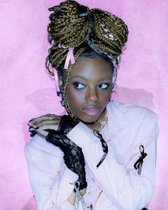  image features Seedel, a poet & mental health advocate, posing against a pink background. With intricate braids & a pink shirt paired with black lace gloves, Seedel radiates style and confidence alongside her contributions to literature & social causees