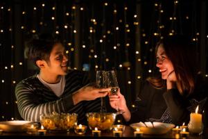 A young couple in black attire celebrates at a candle-lit dinner, toasting their glasses amid a romantic backdrop of twinkling fairy lights.
