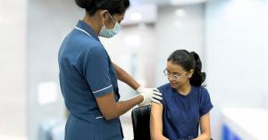 A healthcare professional in a blue uniform and face mask administers a vaccination to a young woman wearing glasses and a blue shirt, seated in a clinic setting.
