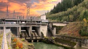A large concrete hydroelectric dam, the Cowlitz Falls Dam, with power lines above, is situated in a forested valley. A calm river flows through it under a vibrant orange and pink sunset sky.
