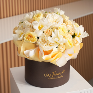 A flower box of peach spray roses, white garden roses and white tulips in a brown colored round box on a white table.