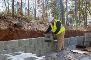 A hardscaping contractor in full safety gear is installing a retaining concrete block wall on a new property surrounded by tall trees, showcasing professional retaining wall services.