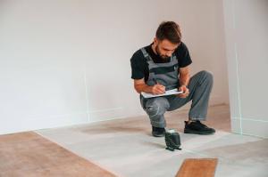 A bearded male contractor wearing a black shirt under denim overalls uses a laser leveler to take measurements and make notes for insurance estimates in a document.