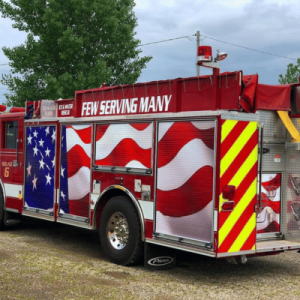 A fire truck decorated with American flag graphics and the phrase "Few Serving Many" is parked on a gravel surface.