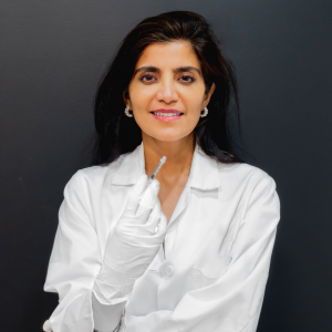 Dr. Lara Devgan holds a medical syringe, smiling against a plain dark background in a white lab coat and gloves.