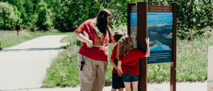 A woman and two children standing together, looking at a sign in a park.