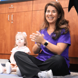 A woman in purple scrubs claps while sitting on the floor next to a young child at St. Tammany Parenting Center.
