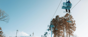 A skier sitting on a ski lift, surrounded by snowy mountains and trees.