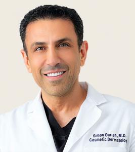 Smiling portrait of Dr. Simon Ourian, M.D., Cosmetic Dermatology expert, wearing a white coat with his name and title embroidered on it.