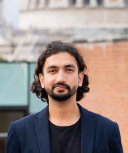 Portrait of Abhinav Jain, a man with medium-length wavy black hair and a beard, wearing a dark blazer over a black shirt. He is standing outdoors with a blurred background featuring a building with a mix of brick and modern glass elements.