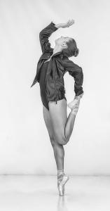 Young female ballet dancer raising her hand into the air as she stands on her right toe holding her left leg in with her left hand.
