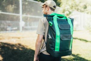 Founder of Team Pickleball, Blake, wearing the RePlay Pickleball Backpack in the color Fern, standing near a fenced pickleball court on a sunny day.