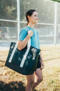 Founder of Team Pickleball, Melanie, wearing the RePlay Pickleball Bag in the color Moon, standing near a fenced pickleball court on a sunny day.