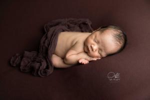 Newborn Boy folded over with chin on feet. Womb Pose. Brown wrap and background