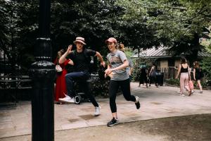 A young man in a white bucket hat and a woman in a pink baseball cap run smiling down a New York City sidewalk while participating in a large puzzle adventure.