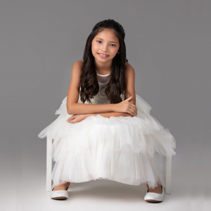  A young girl in a white dress sits gracefully on a white chair, exuding innocence and tranquility in a serene setting.