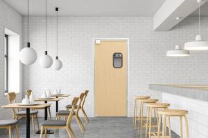 A minimalist café interior featuring wooden tables, chairs, and stools. Pendant lights hang from the ceiling, and a closed wooden door with a small window is in the center of the white brick wall.