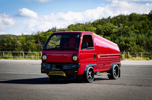 Custom Honda Acty mini truck in vibrant red with aftermarket body kit, featuring sleek black wheels and modern LED headlights, parked outdoors with a scenic forest backdrop.