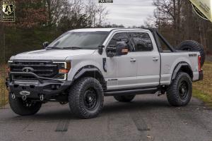 A white MASTIFF® truck equipped with advanced armoring technology, large tires, and a roll bar is parked on a grey road in a wooded area.