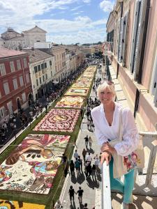 This is a photo of Denise Kowal, the founder of the Sarasota Int'l Chalk Festival on a balcony overlooking the flower carpets made during the 2024 Genzano di Roma Infiorata. The flower carpets are all natural flowers with the use of grains, seeds and eart