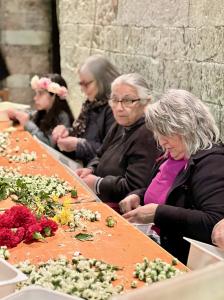 Three women and a girl are sitting at a table with flower on the table, they are pulling the petals off the flowers and sorting them for artists to use in their infiorata paintings in Spello Italy. The walls behind them are old stone, in the historic cent