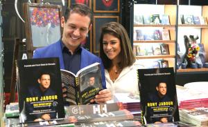 Rony Jabour and his wife holding his newly launched book at the Brazil International Book Fair, both smiling and standing in front of a booth with books displayed.