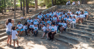 A group of around 40 young kids and camp counselors posing for a group photo all wearing white t-shirts with a Jay Nolan Camp logo on them.